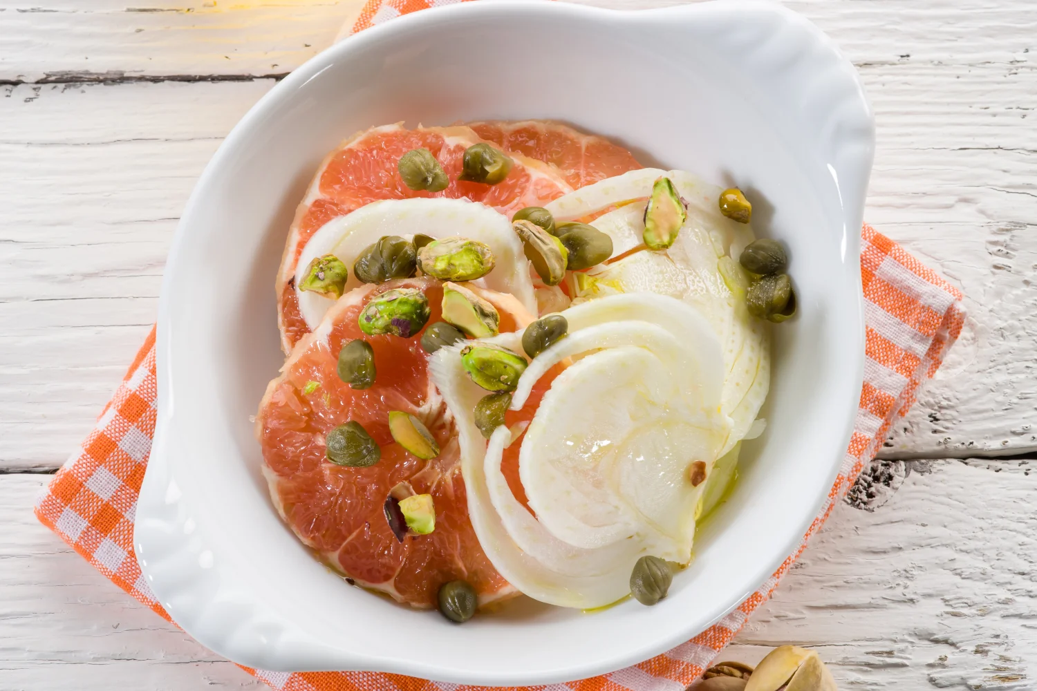 A serving platter of thinly sliced fennel and grapefruit segments scattered with pistachios, capers and fresh herbs, drizzled with a light citrus dressing