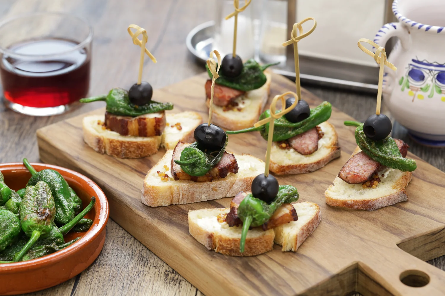 A Spanish pintxo of garlic toast topped with a slice of crispy tocino, a blistered green pepper and a black olive, skewered with a toothpick