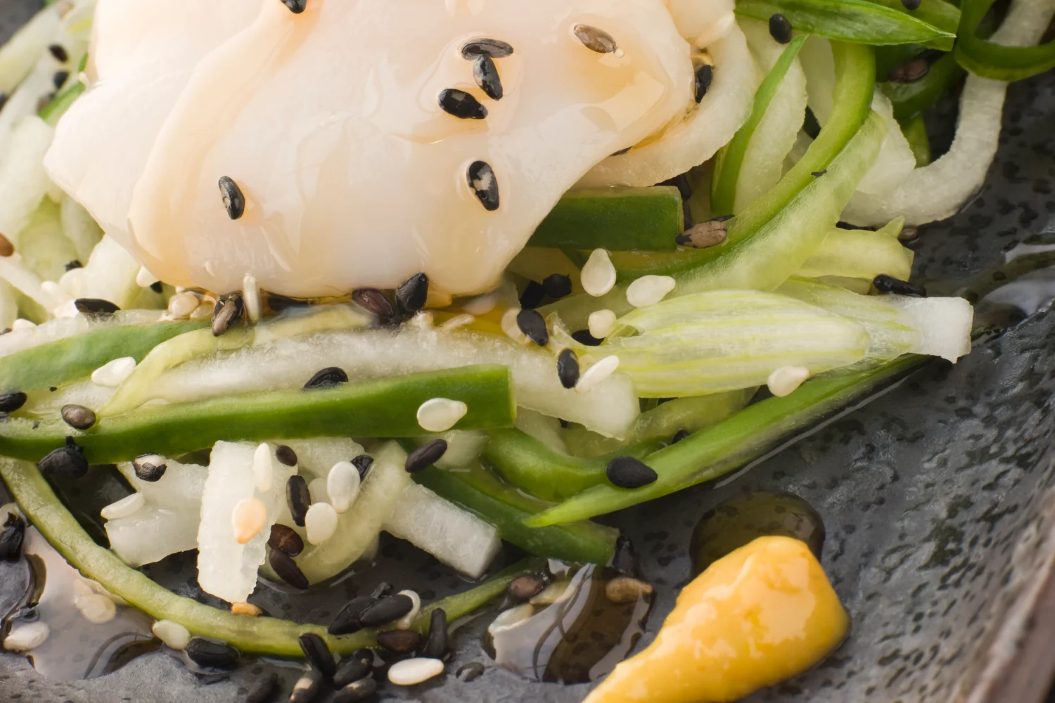 A chilled plate of thinly sliced raw scallops arranged around cucumber julienne, dotted with a yellow turmeric and Dijon dressing and finished with black and white sesame seeds