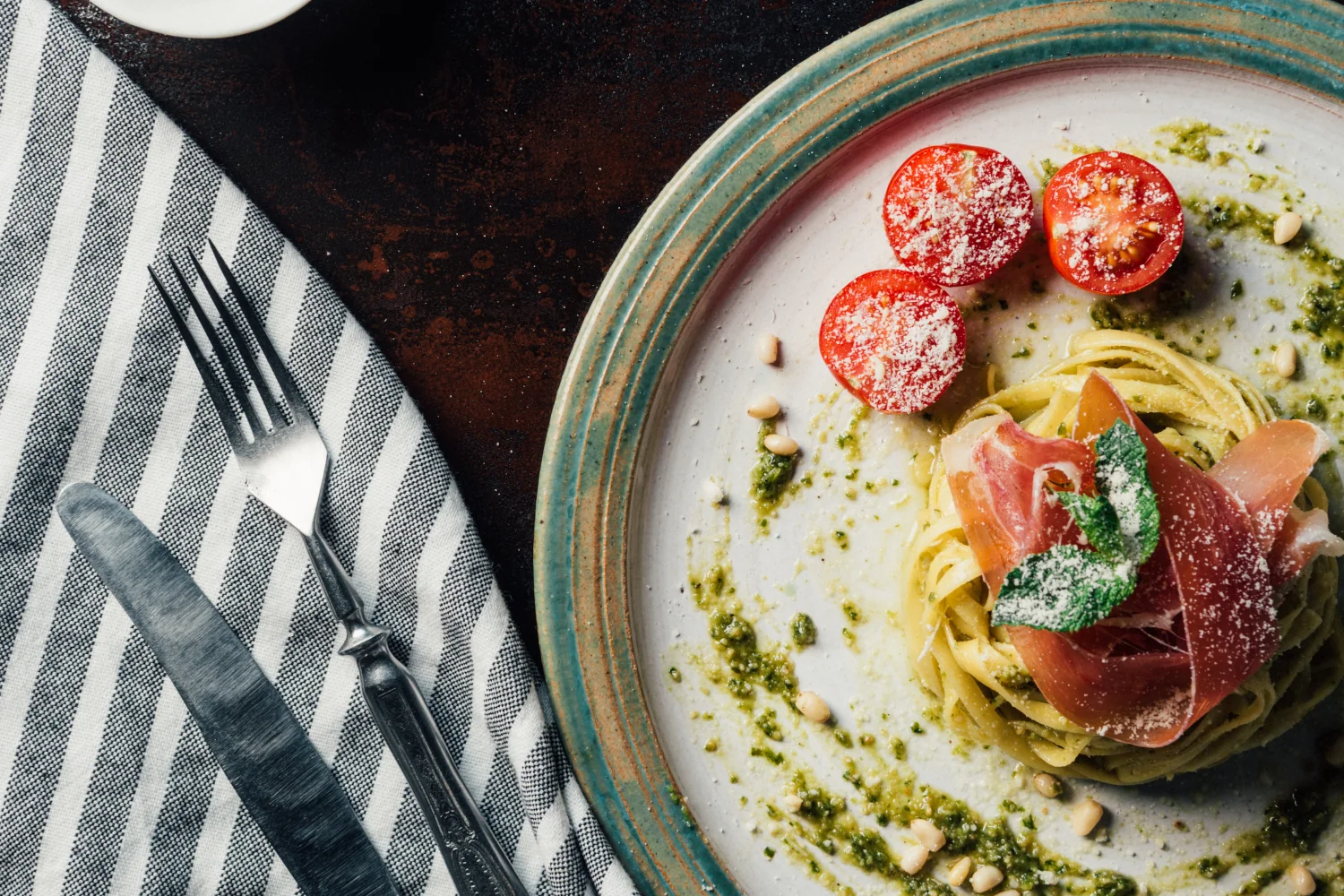 A nest of tagliolini drizzled with vibrant green pesto, topped with strips of raw Parma ham, halved cherry tomatoes, pine nuts and freshly grated Parmesan