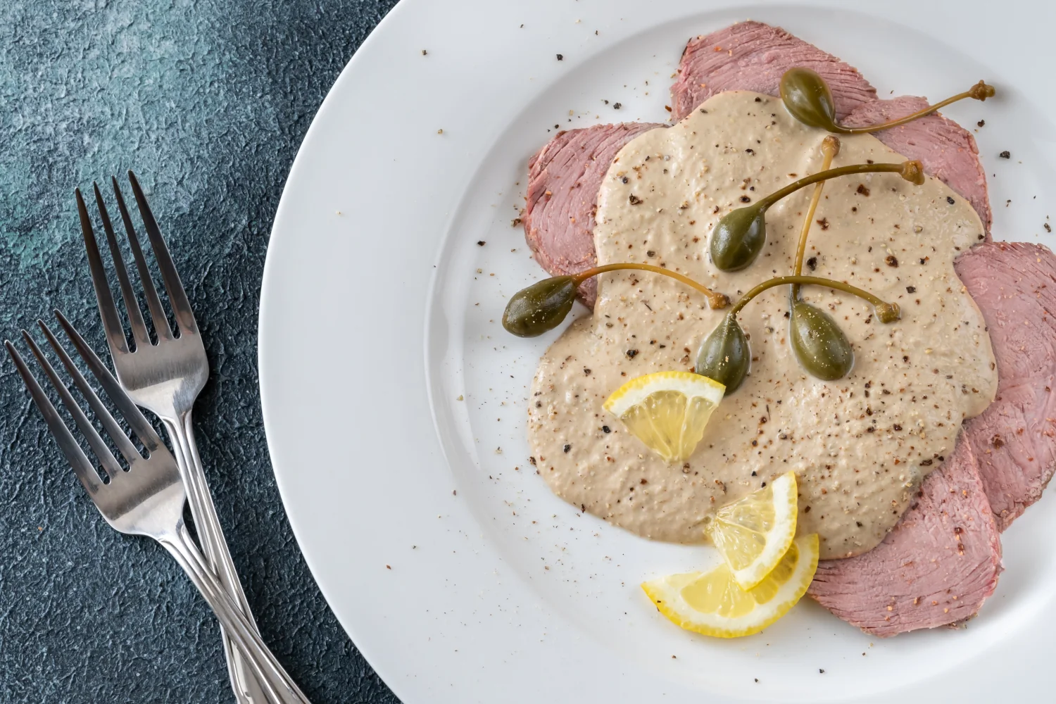 Vitello Tonnato: A serving platter of thinly sliced poached veal covered in a smooth, creamy tonnato sauce of tuna, anchovies and capers, garnished with capers and lemon slices
