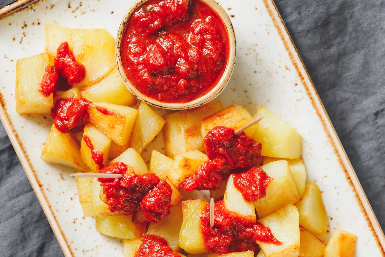 A bowl of double-fried crispy patatas bravas generously topped with a rich, deep-red salsa brava made with smoked pimentón and sherry vinegar